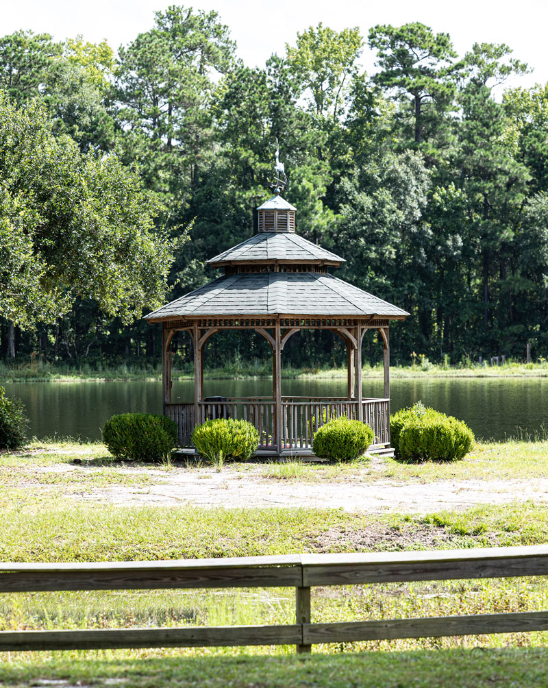 A wooden gazebo stands near a pond, surrounded by green trees and bushes, with a wooden fence in the foreground on a sunny day.