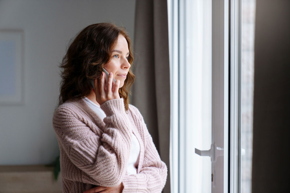 Woman with wavy brown hair wearing a pink cardigan stands by a window, looking outside thoughtfully with her hand resting on her cheek.
