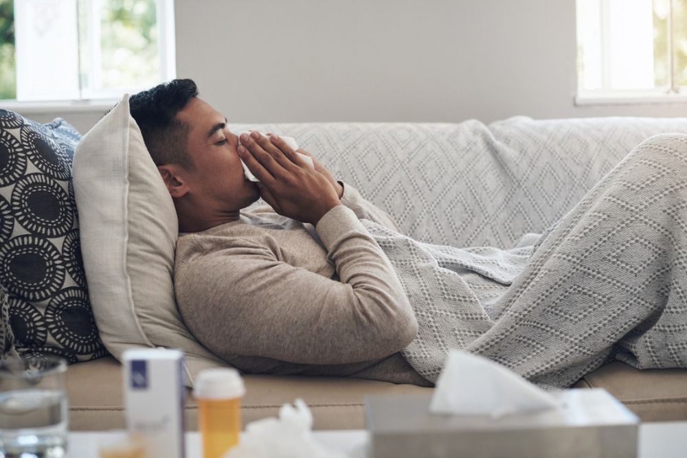 A person lying on a couch under a blanket, holding a tissue to their face. Medicine, tissues, and a glass of water are on the table nearby, suggesting they are unwell.
