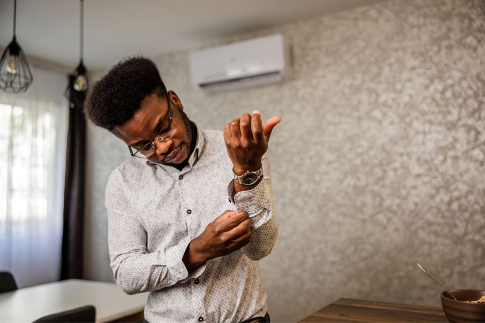 A man wearing glasses and a patterned shirt stands by a table while adjusting his sleeve. A bowl and spoon are on the table, and light enters through a window in the background.