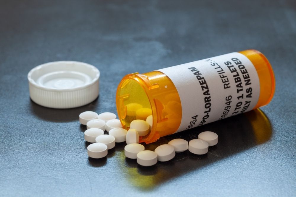 An orange prescription pill bottle labeled Lorazepam tipped over with white round tablets spilling out, next to the bottles white cap, on a gray surface.