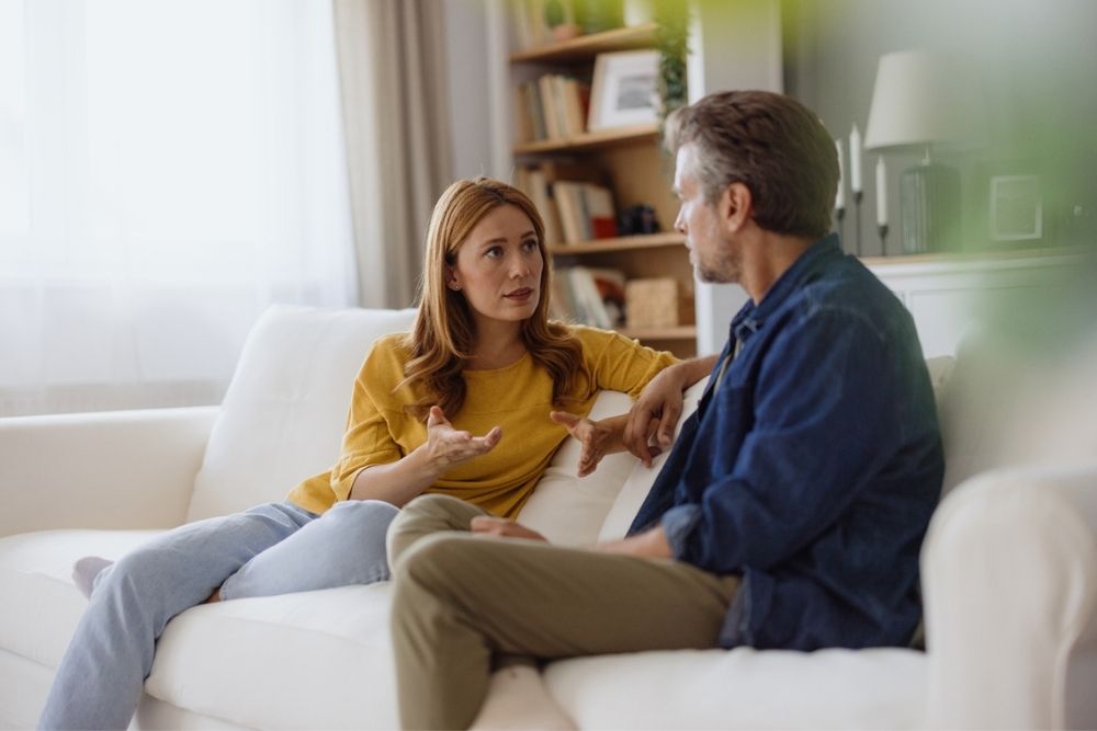 A woman and a man sit on a white couch in a living room, engaged in a serious conversation. The woman gestures with her hands while speaking, and the man listens attentively.