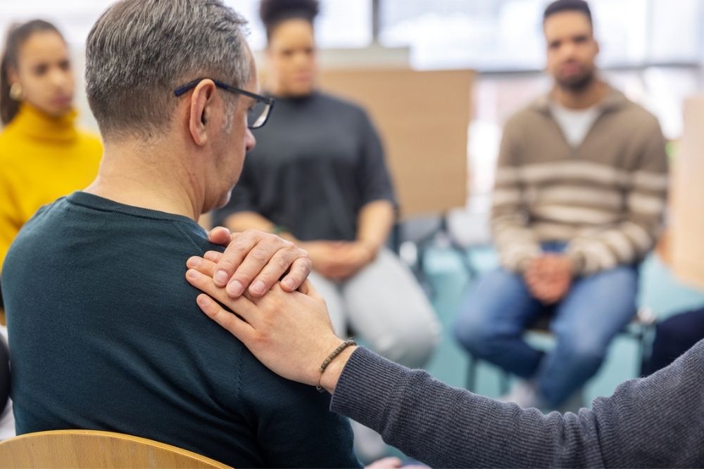 A person comforts another by placing a hand on their shoulder during a group therapy session, with several people sitting in a circle in the background.