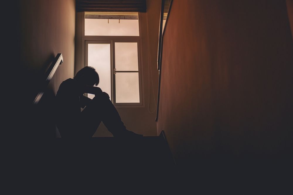 A person sits alone on a dimly lit staircase, knees up and head resting on their arms, silhouetted against a window with soft light coming through.