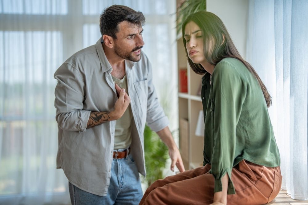 A man appears to be arguing with a woman who looks upset and is avoiding eye contact while sitting on a table. Sunlight streams through windows in the background, and there are plants visible.