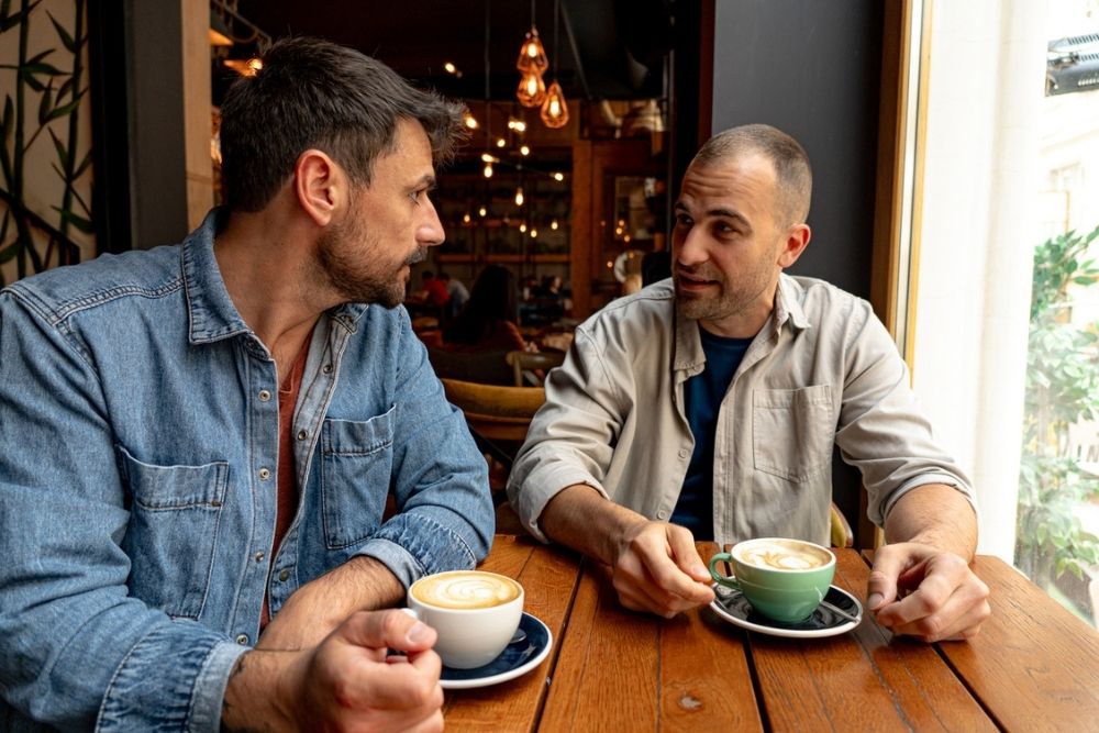 Two men sit at a wooden table in a café, each with a cup of coffee, engaged in conversation while natural light streams through the window beside them.