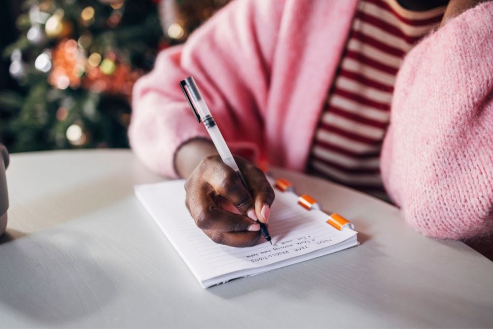 A person wearing a pink sweater and striped shirt writes in a notebook with a pen at a table. A blurred Christmas tree is visible in the background.