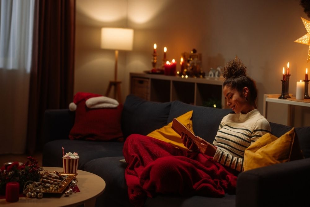 A woman sits on a couch reading a book, wrapped in a red blanket, with festive holiday decorations, candles, and warm lighting creating a cozy atmosphere.