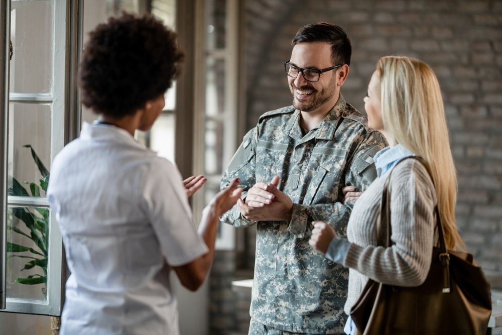 Smiling person in military uniform stands with a woman, talking to another woman indoors. All appear happy and engaged in conversation.