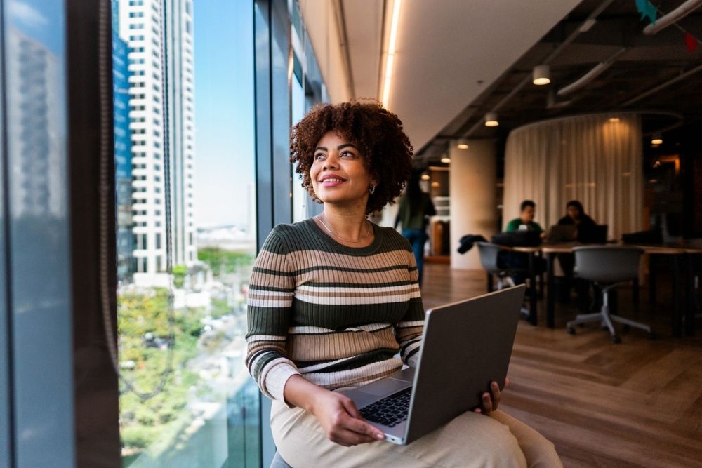 A woman sits by a large window in a modern office, holding a laptop and smiling while looking outside. Tall buildings and a cityscape are visible through the window.