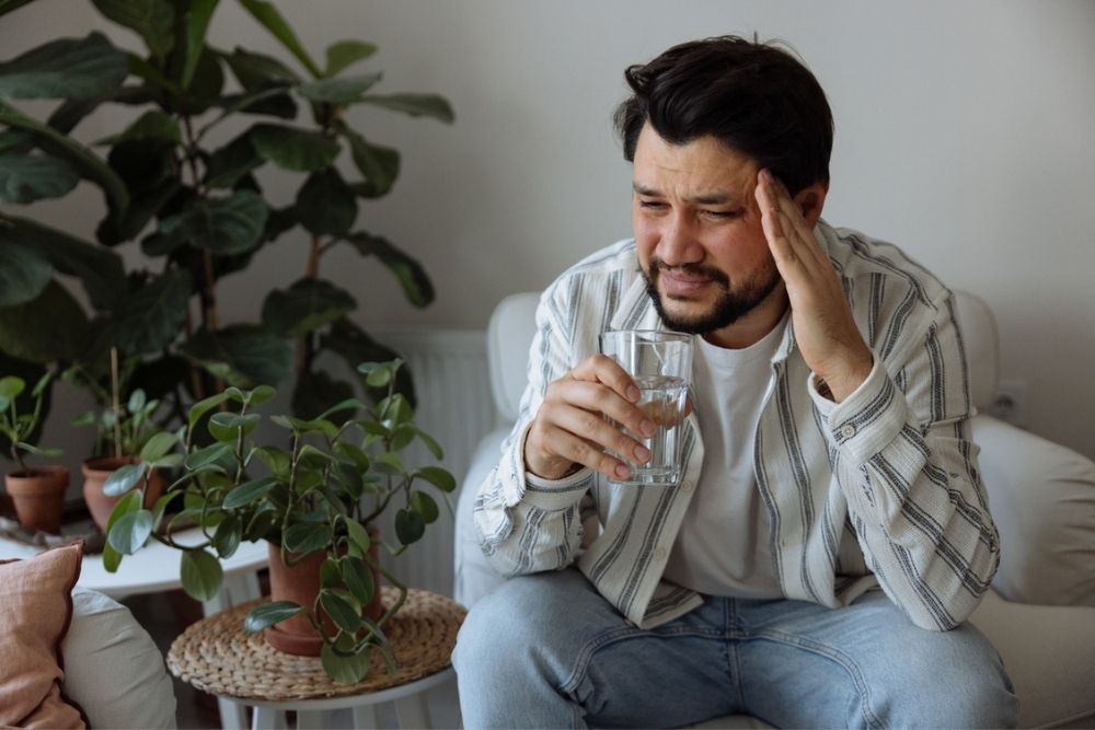 A man sits on a couch holding a glass of water and touching his head, looking like he has a headache. There are green potted plants around him.