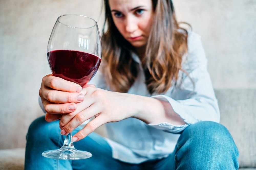 A woman in a white shirt and jeans sits holding a large glass of red wine, looking intently at the drink with a serious expression.