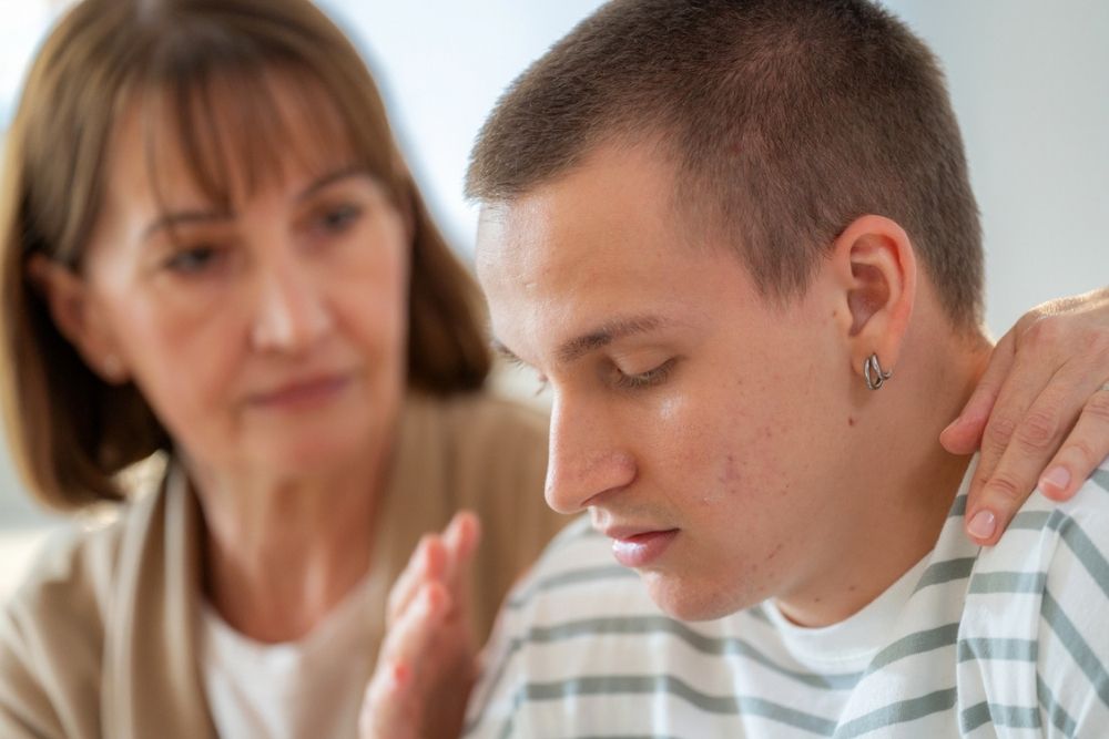 A woman gently touches the shoulder of a young man who appears upset or deep in thought, offering comfort and support.
