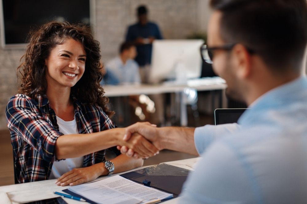 A woman and a man sit across a desk, smiling and shaking hands. Papers and a clipboard are on the desk. Other people work in the background in a modern office setting.