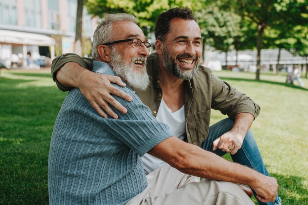 Two men, one older and one younger, sit on grass outdoors, smiling and hugging each other in a park on a sunny day.