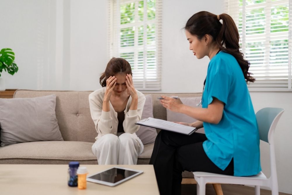 A woman sits on a couch holding her head in distress while a healthcare professional in scrubs talks to her, holding a clipboard. A table with a tablet and medication bottles is in the foreground.