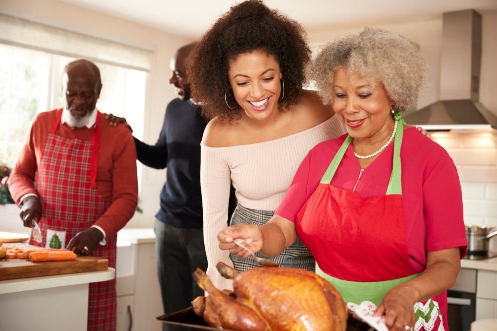 Four people in a kitchen prepare a holiday meal. An older woman holds a roasted turkey, a young woman smiles beside her, while two men in the background chop vegetables and talk.