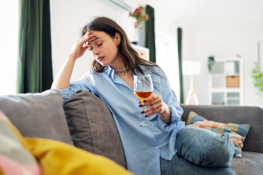 A woman sits on a couch holding a glass of wine, looking down with her hand on her forehead, appearing stressed or tired in a bright living room.