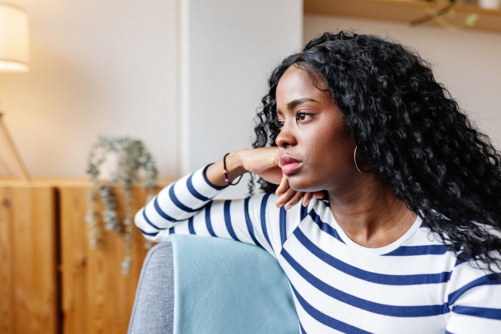 A woman with long curly hair, wearing a striped shirt, sits on a couch looking thoughtfully out of frame, resting her arm on the back of the seat in a bright, modern room.