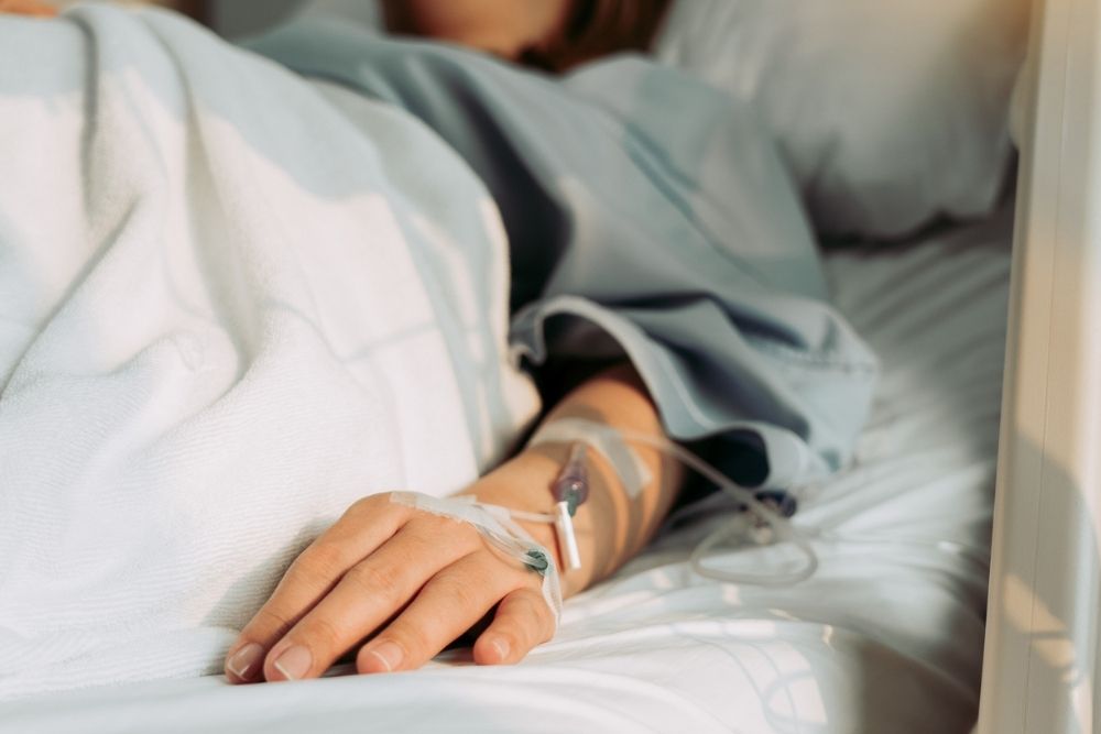 A person lies in a hospital bed with an IV inserted in their hand, covered by a white blanket. Sunlight shines softly over the scene.