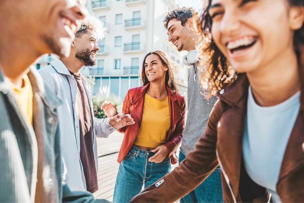 Four young adults stand outside in sunlight, smiling and laughing together. They appear to be friends enjoying a pleasant day in an urban area, dressed in casual clothing.