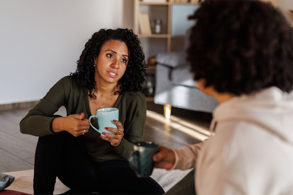 Two people sit indoors, holding mugs and having a serious conversation. One person with curly hair and a green sweater gestures while speaking, while the other listens. Shelves and a sofa are visible in the background.