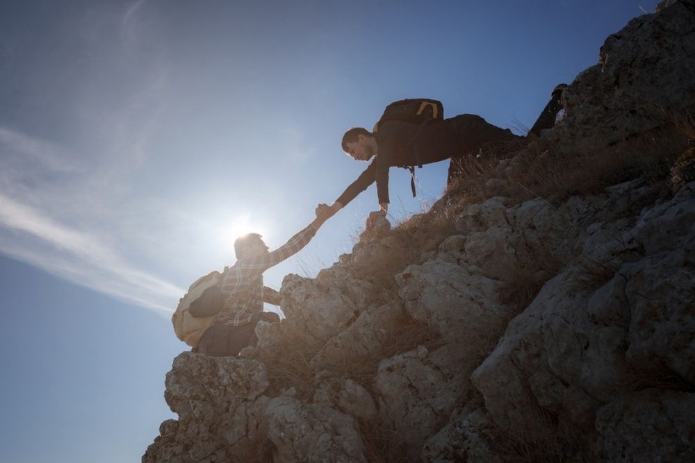 Two hikers on a rocky slope, one reaching down to help the other up. The sun shines brightly behind them, creating a silhouette effect. Both wear backpacks and are climbing outdoors.
