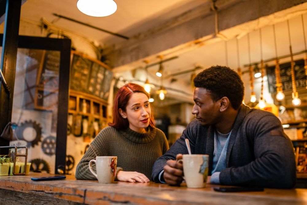 Two people sit at a cozy café counter, engaged in conversation. Both have mugs in front of them. Warm lighting and industrial décor create a relaxed atmosphere.