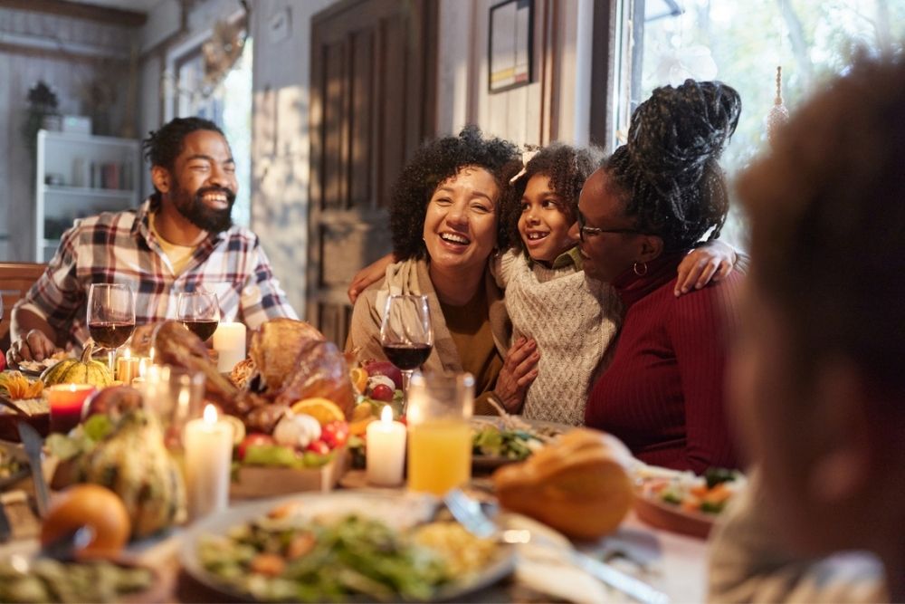 A family sits together at a festive dinner table, smiling and hugging, surrounded by candles, food, and autumn decorations in a cozy, sunlit room.