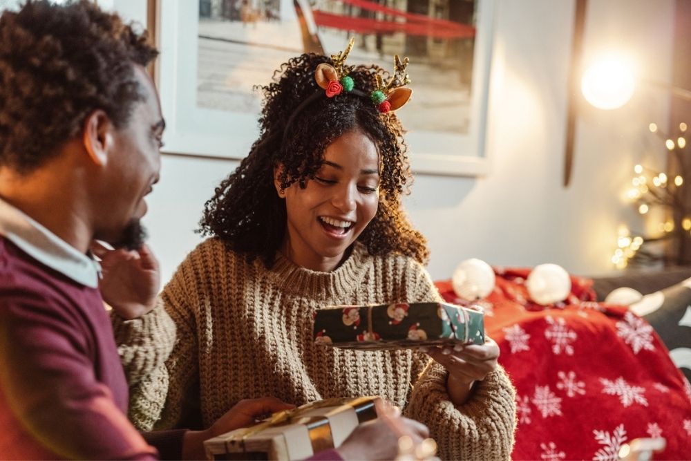 A woman wearing reindeer antlers and a brown sweater smiles as she opens a Christmas gift, sitting next to a man in a cozy, festive living room.