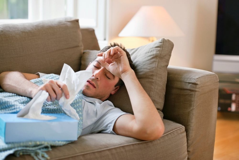 A man lies on a couch under a blanket, holding a tissue and looking unwell. A tissue box is nearby. There is a lamp and window in the background, suggesting he is at home resting.