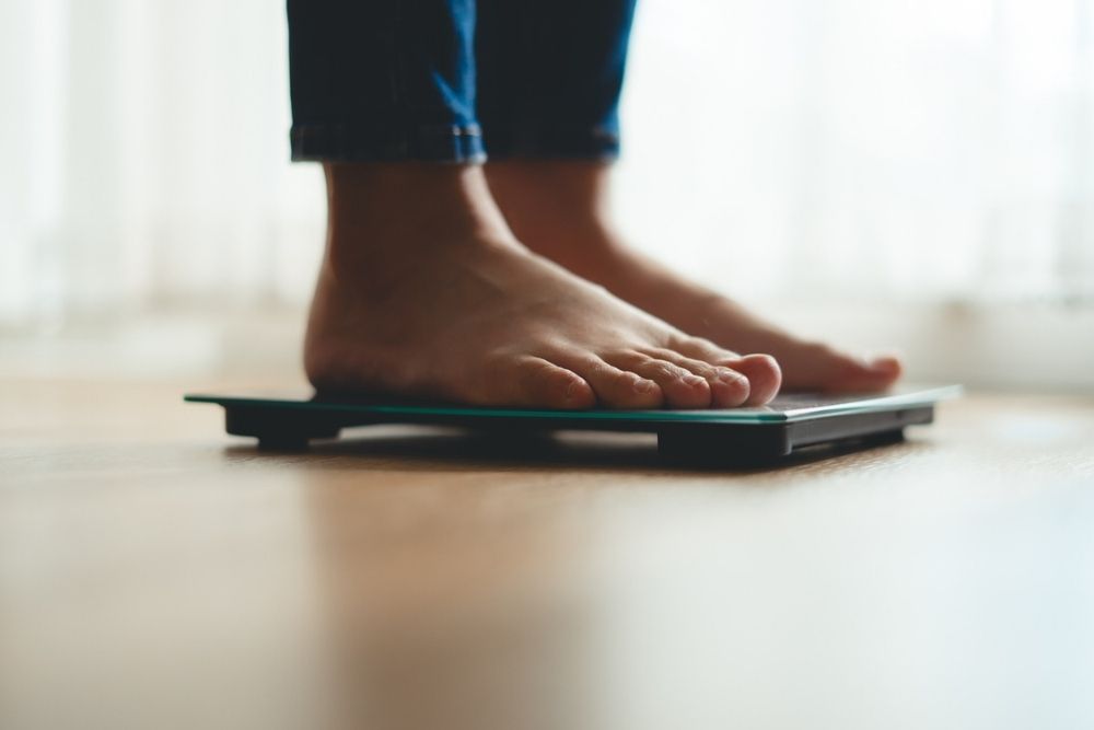 Close-up of a persons bare feet standing on a digital bathroom scale on a wooden floor, with light coming through a sheer curtain in the background.