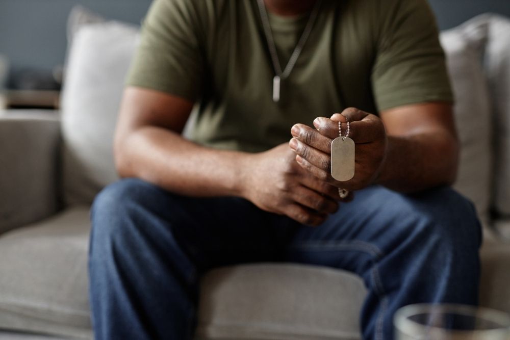 A person in a green shirt and jeans sits on a couch, holding a metal dog tag necklace in their hands. The focus is on the hands and dog tag; the persons face is not visible.