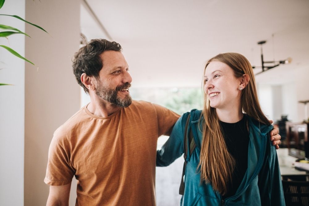 A man and a young woman stand indoors, smiling at each other. The man has his arm around the womans shoulders and they both look happy.