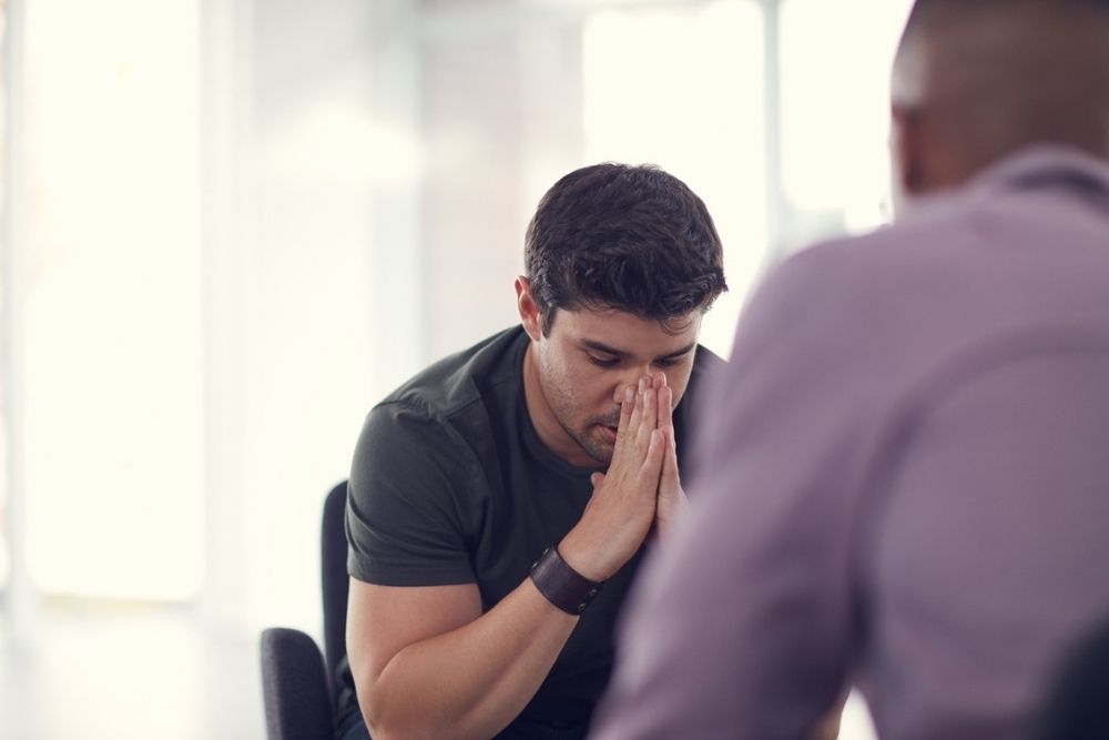 A man sits with his hands pressed to his face, looking stressed, while another person sits across from him, partially visible and blurred in the foreground.