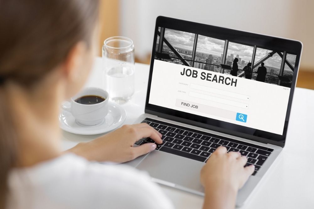 A person using a laptop with a Job Search website open, typing on the keyboard. A cup of coffee sits nearby on the table.