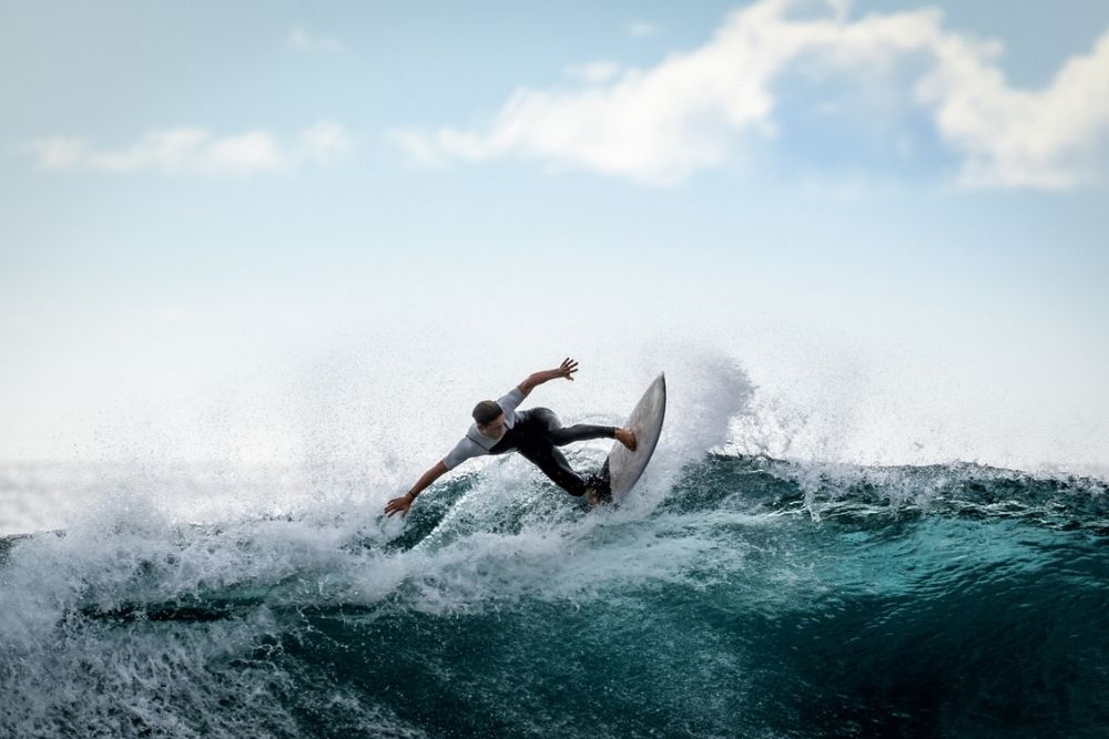 A surfer in a white shirt rides a wave, balancing skillfully on a surfboard with arms outstretched. The ocean wave is splashing, and the sky is bright with some clouds.