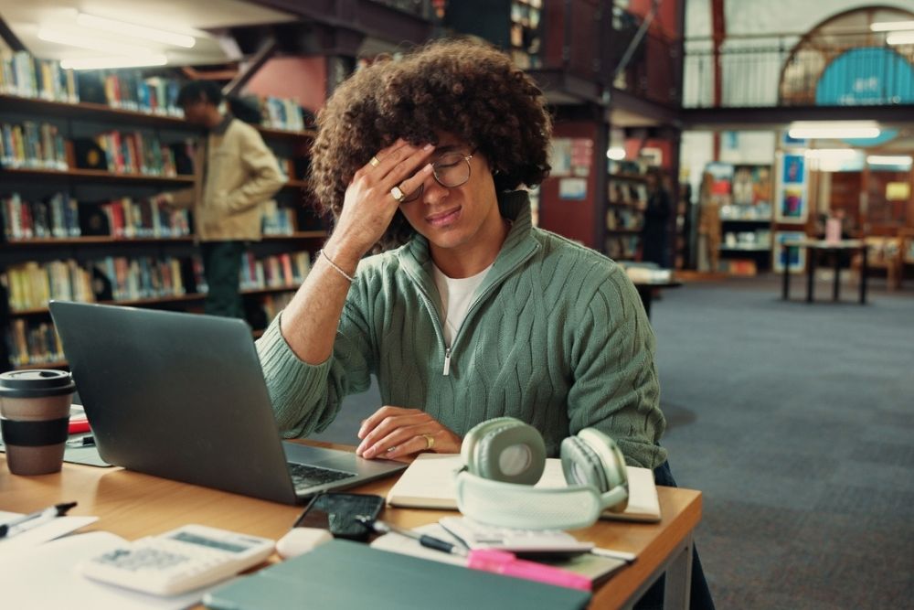 A person sits at a desk in a library, eyes closed and hand on forehead, looking stressed. An open laptop, books, coffee cup, and headphones are on the desk. Bookshelves and another person are in the background.