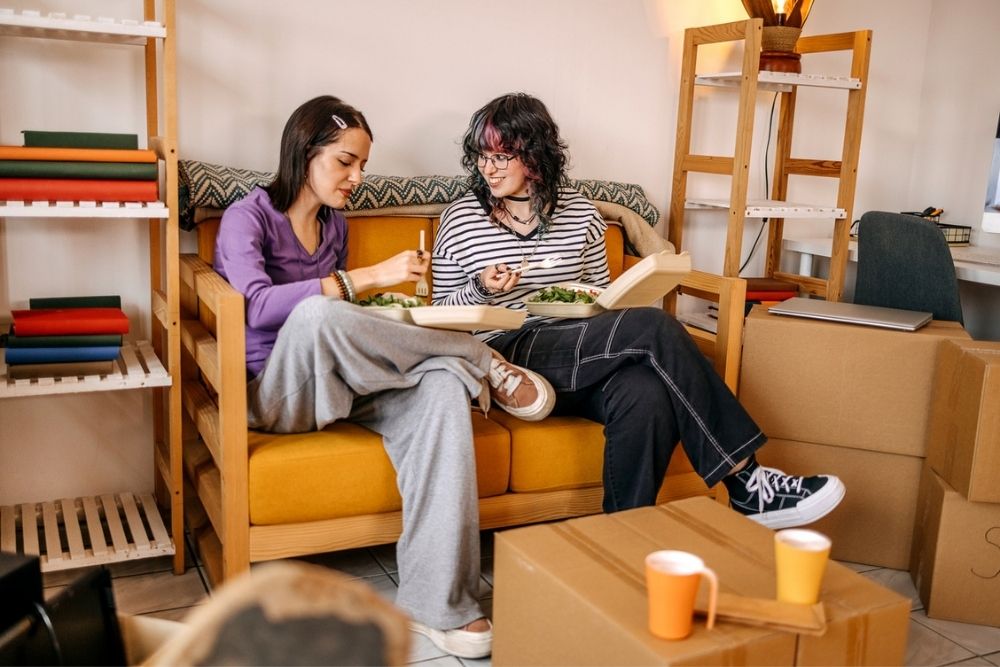 Two people sit on a yellow couch eating takeout food, surrounded by moving boxes and bookshelves, suggesting they are in the process of moving into a new home.