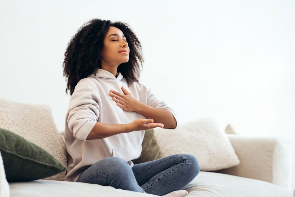 Young woman with curly hair sits cross-legged on a couch, eyes closed, one hand on her chest and the other on her lap, appearing calm and relaxed, possibly meditating or practicing deep breathing.