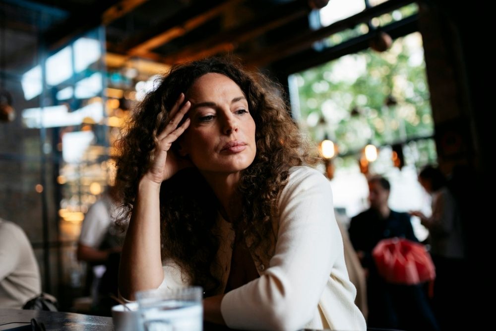A woman with curly hair sits at a table in a cafe, resting her head on her hand and looking thoughtful. There is a glass of water in the foreground and blurred people in the background.
