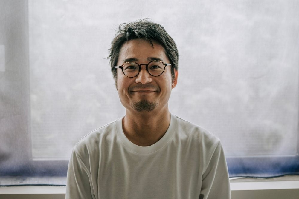 A man with short dark hair, glasses, and a trimmed beard smiles gently while wearing a white t-shirt, sitting indoors in front of a light-colored, softly lit window.