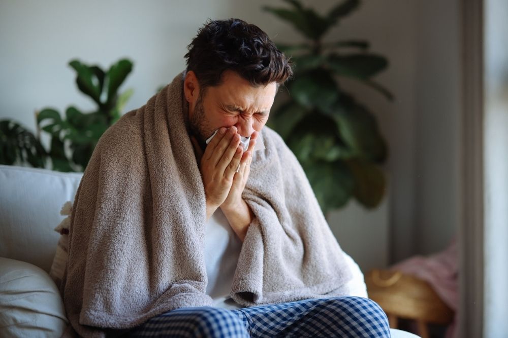 A man sits on a couch wrapped in a blanket, sneezing into a tissue. He appears unwell or sick, with indoor plants visible in the background.