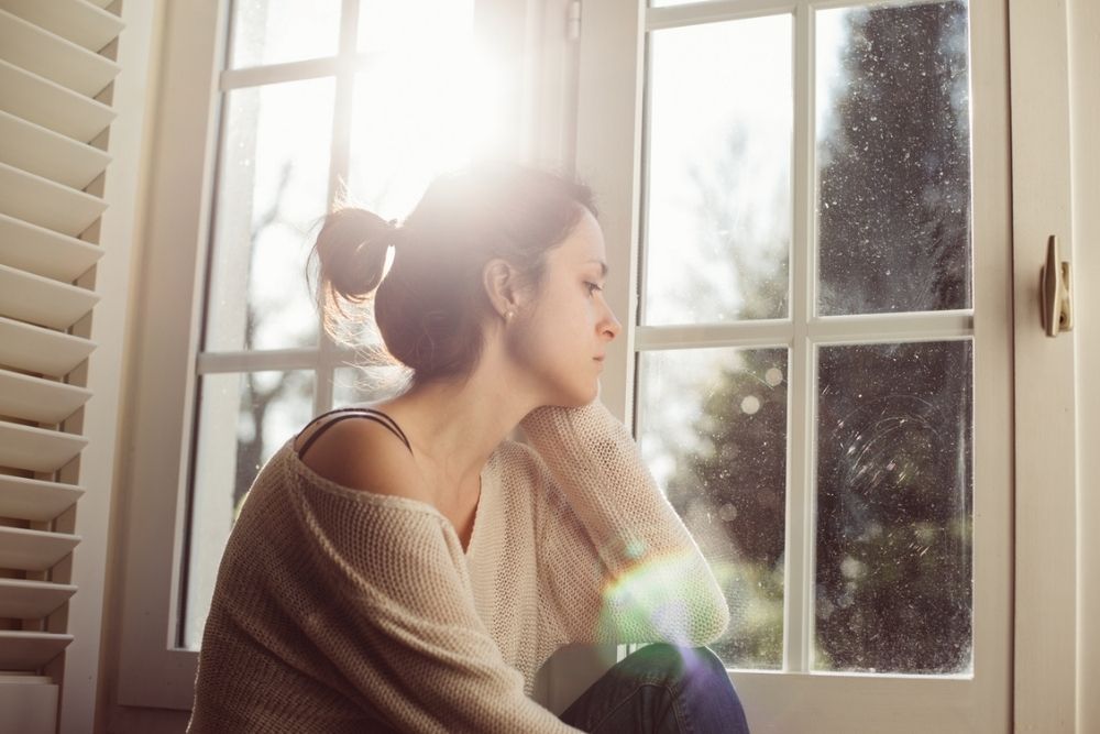 A woman with her hair in a bun sits by a window with sunlight streaming in, resting her head on her hand and gazing outside with a thoughtful expression.