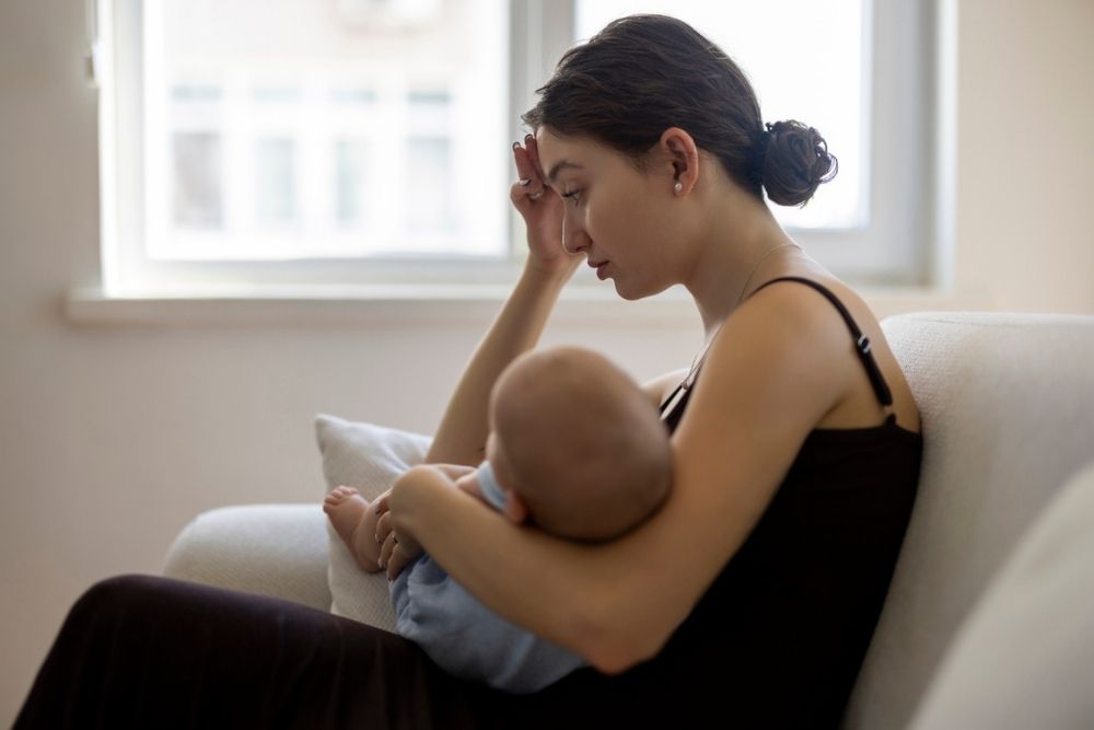 A woman sits on a couch, breastfeeding a baby. She looks tired or stressed, with her hand on her forehead, gazing downward. A window is in the background, and light fills the room.