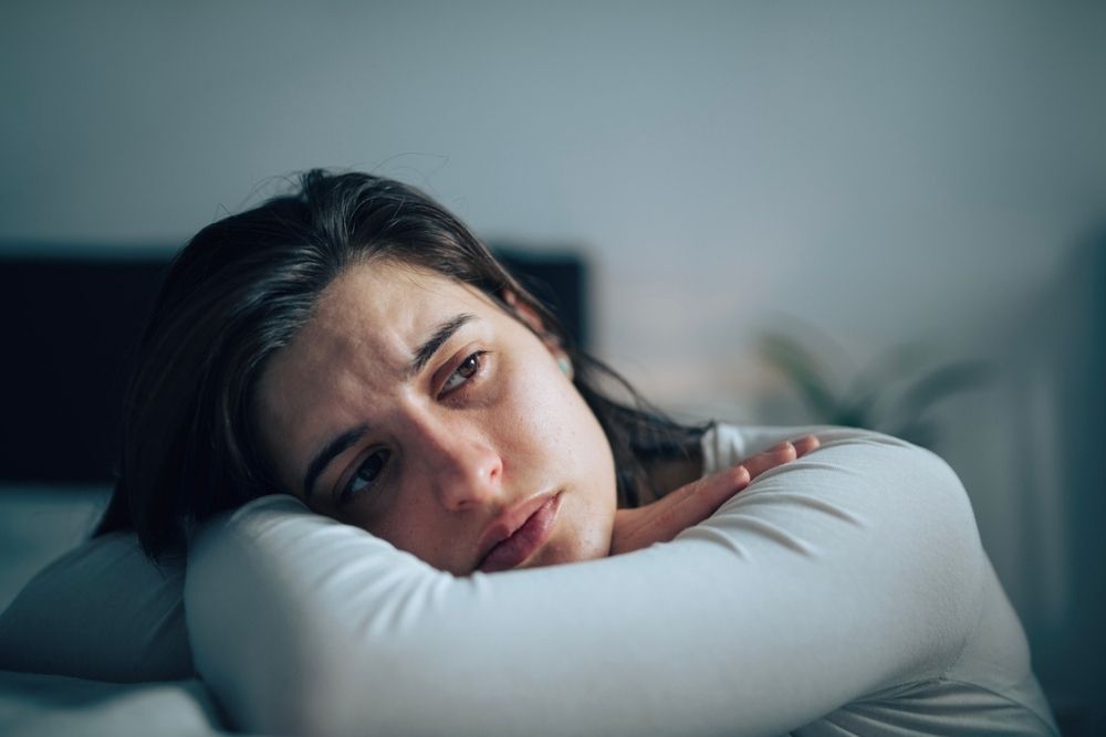 A woman with long brown hair rests her head on her arms, looking away with a sad, thoughtful expression. The background is blurred, suggesting an indoor setting.