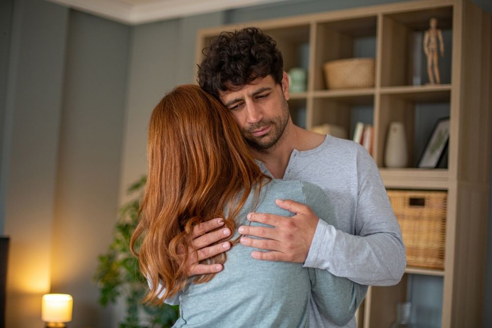 A man and a woman hug in a living room. The man appears concerned or sympathetic while holding the woman close, offering comfort. Shelves with books and decor are visible in the background.