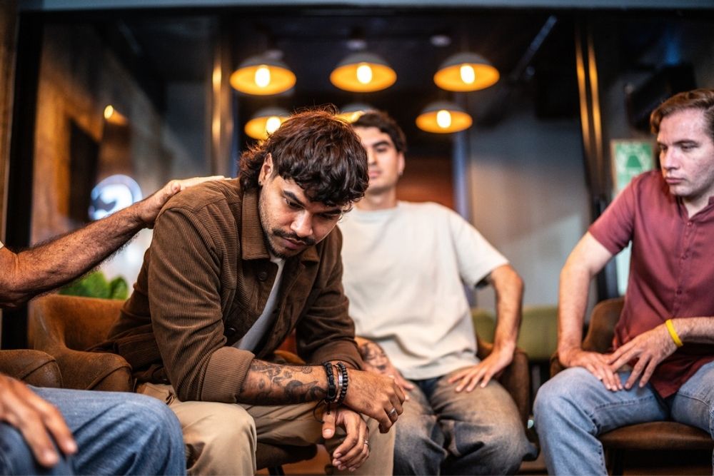 A man sits with his head down, looking distressed, while others around him offer comfort and support, with one person resting a hand on his shoulder.