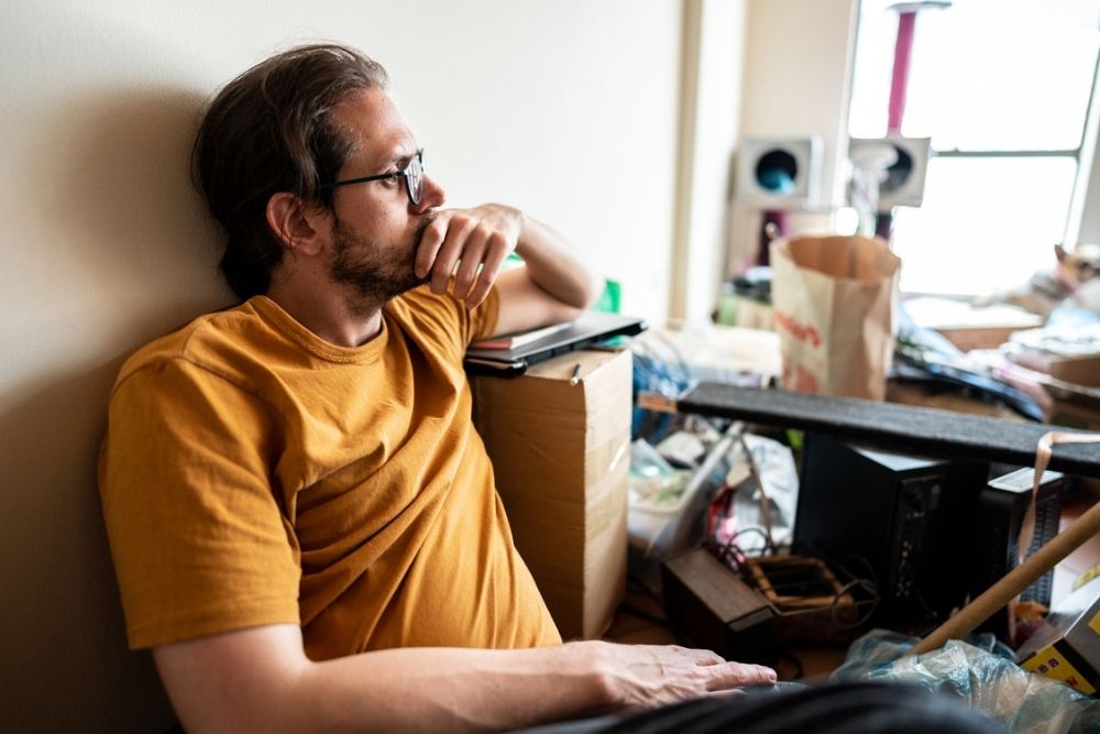 A man in glasses and a yellow shirt sits on the floor, looking thoughtful, surrounded by clutter and boxes in a messy room with light coming through a nearby window.