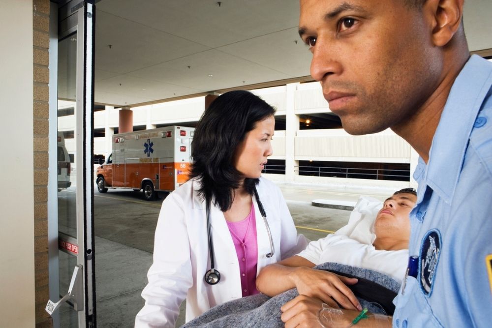 A doctor and paramedic accompany a patient on a stretcher into a hospital, with an ambulance parked outside the entrance.